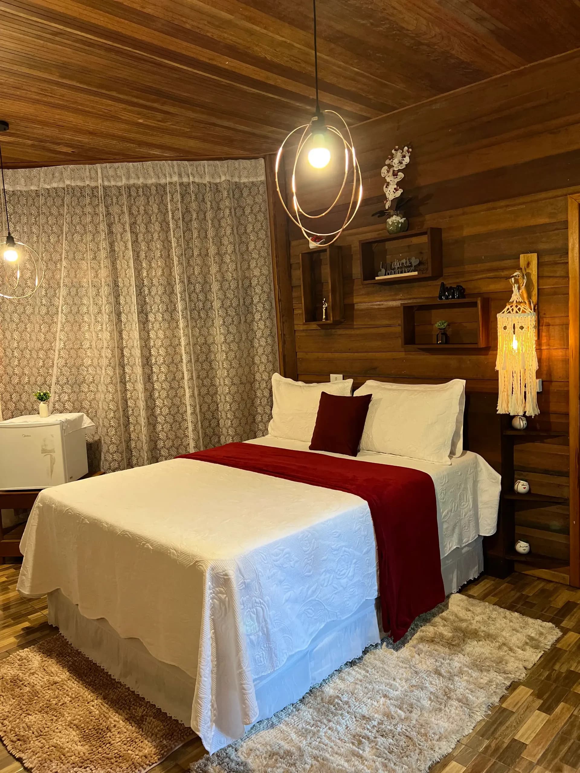 Quarto com paredes de madeira, cama com manta vinho e luminárias pendentes