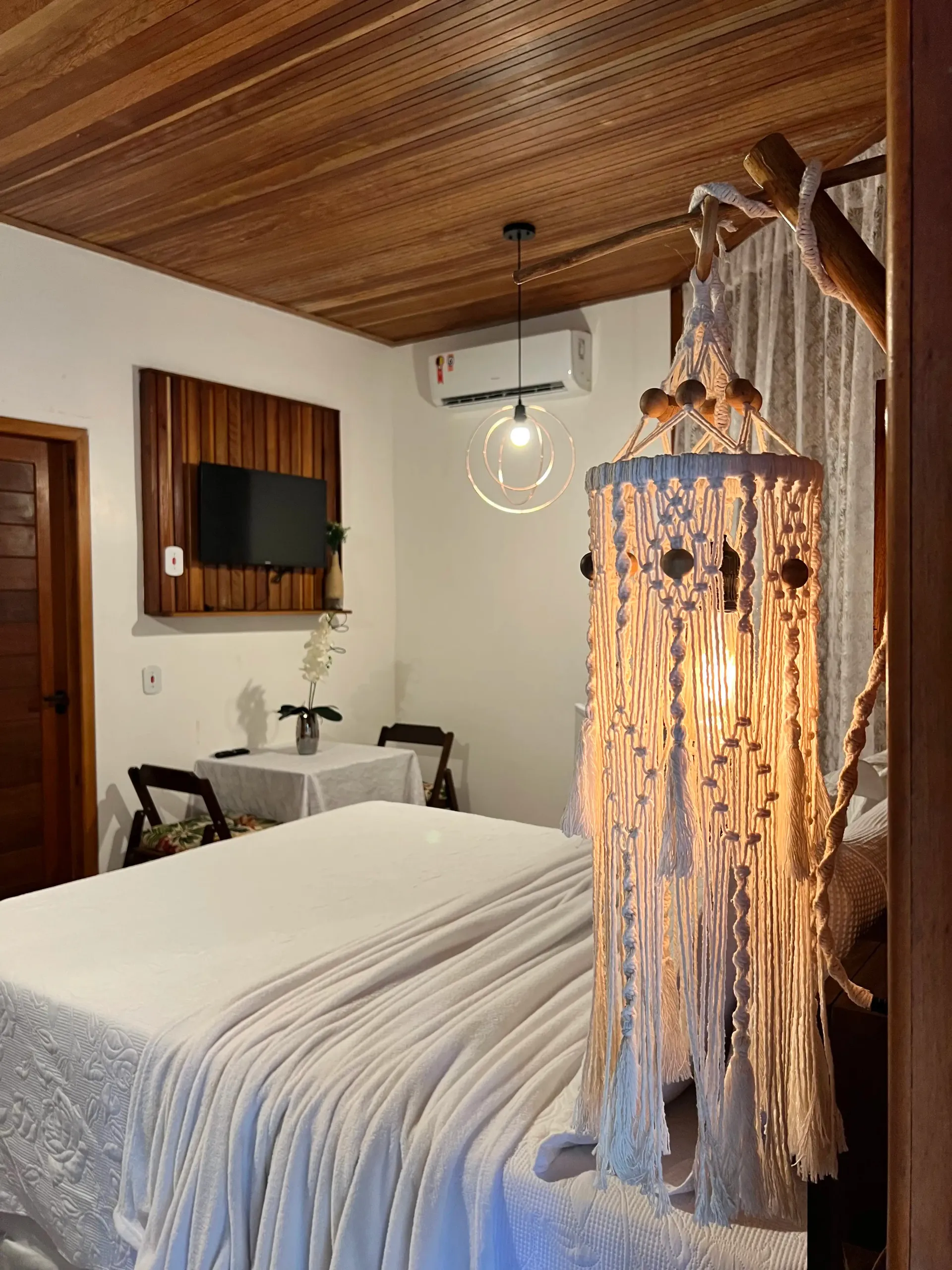 Quarto com forro de madeira, cama de enxoval branco e luminária de macramê acesa