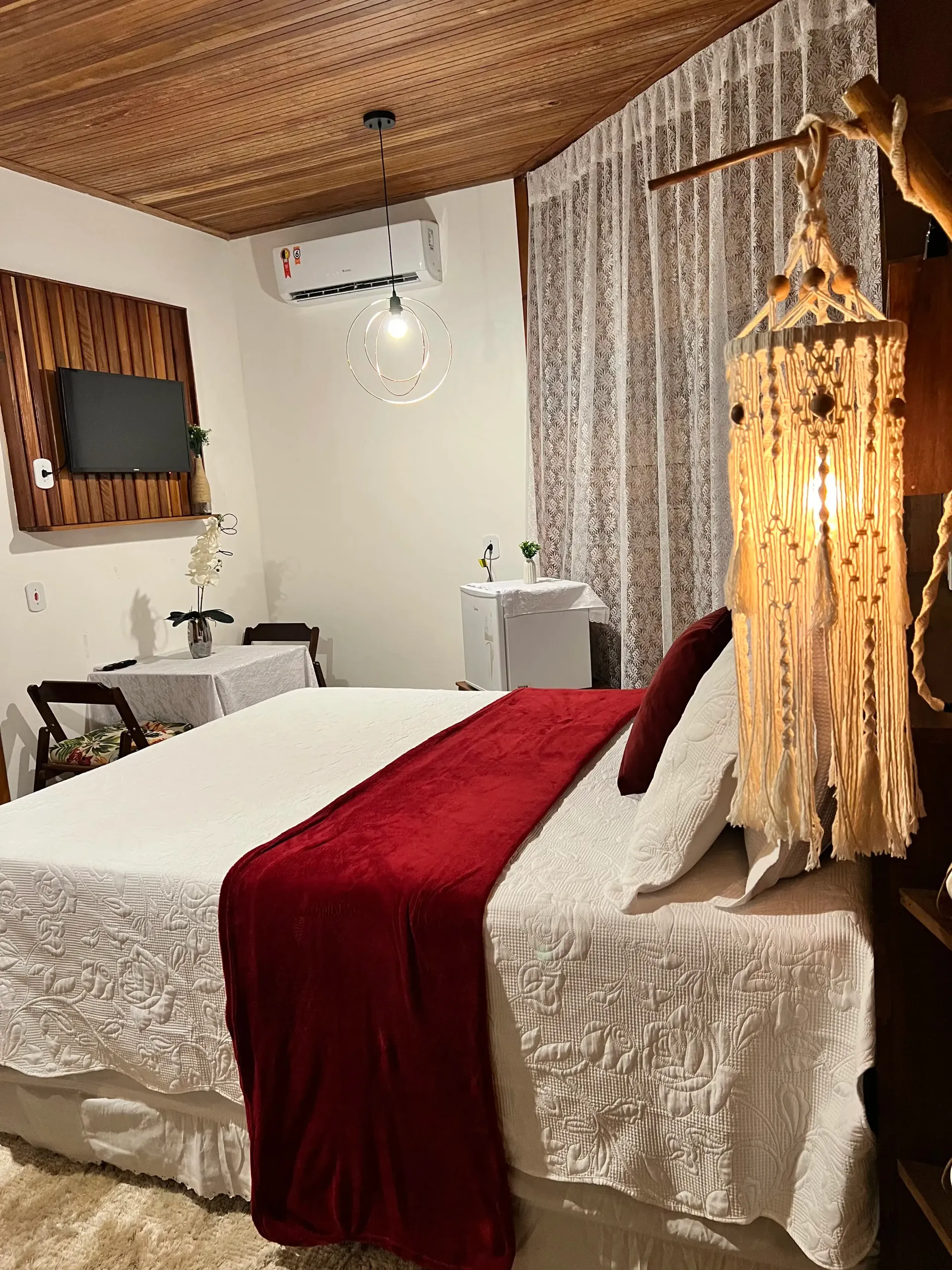 Cama com manta vermelha e luminária de macramê em quarto com forro de madeira