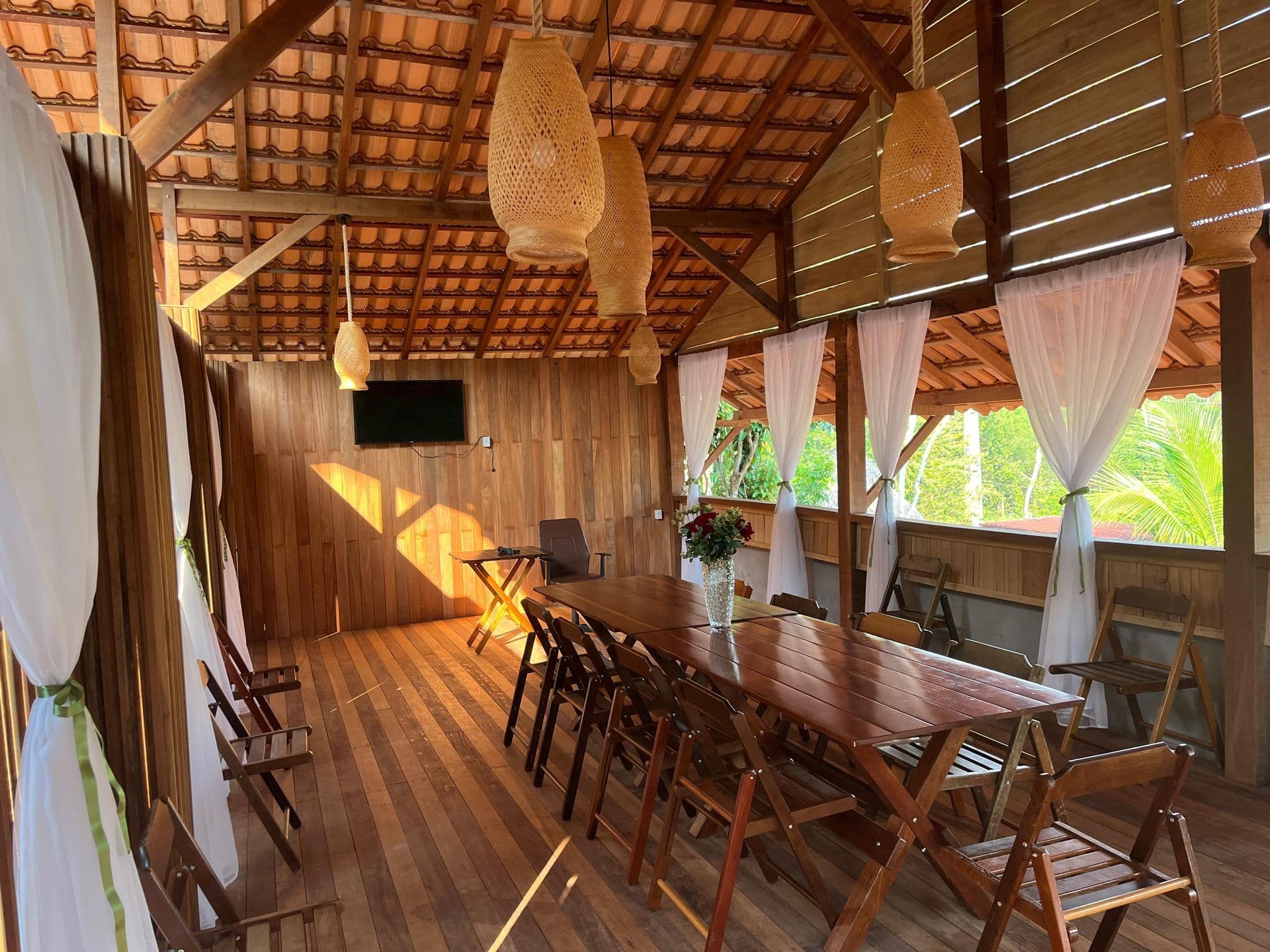 Deck de madeira com mesa longa e luminárias de fibra penduradas, cortinas brancas nas laterais
