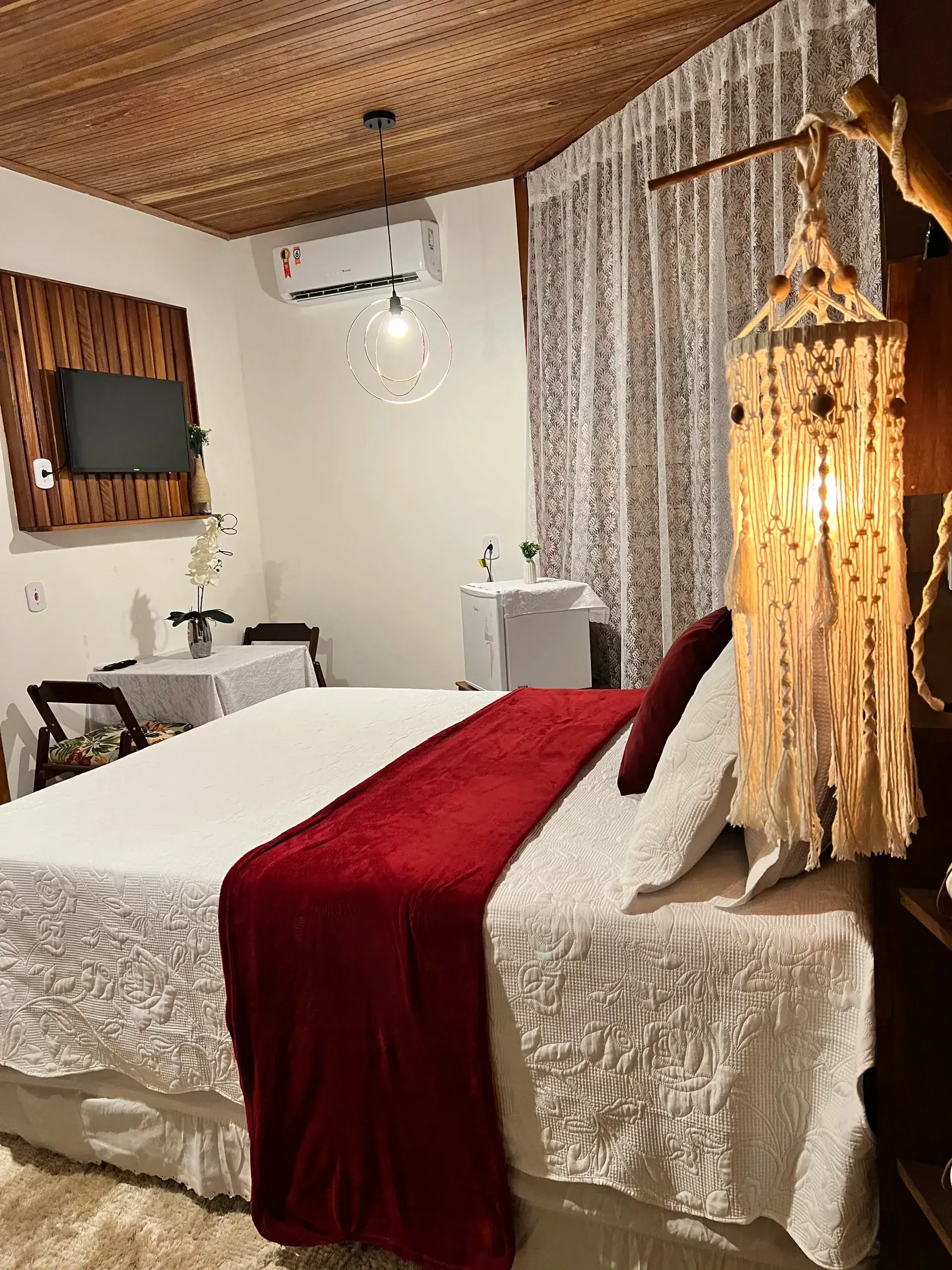 Cama com manta vermelha e luminária de macramê em quarto com forro de madeira
