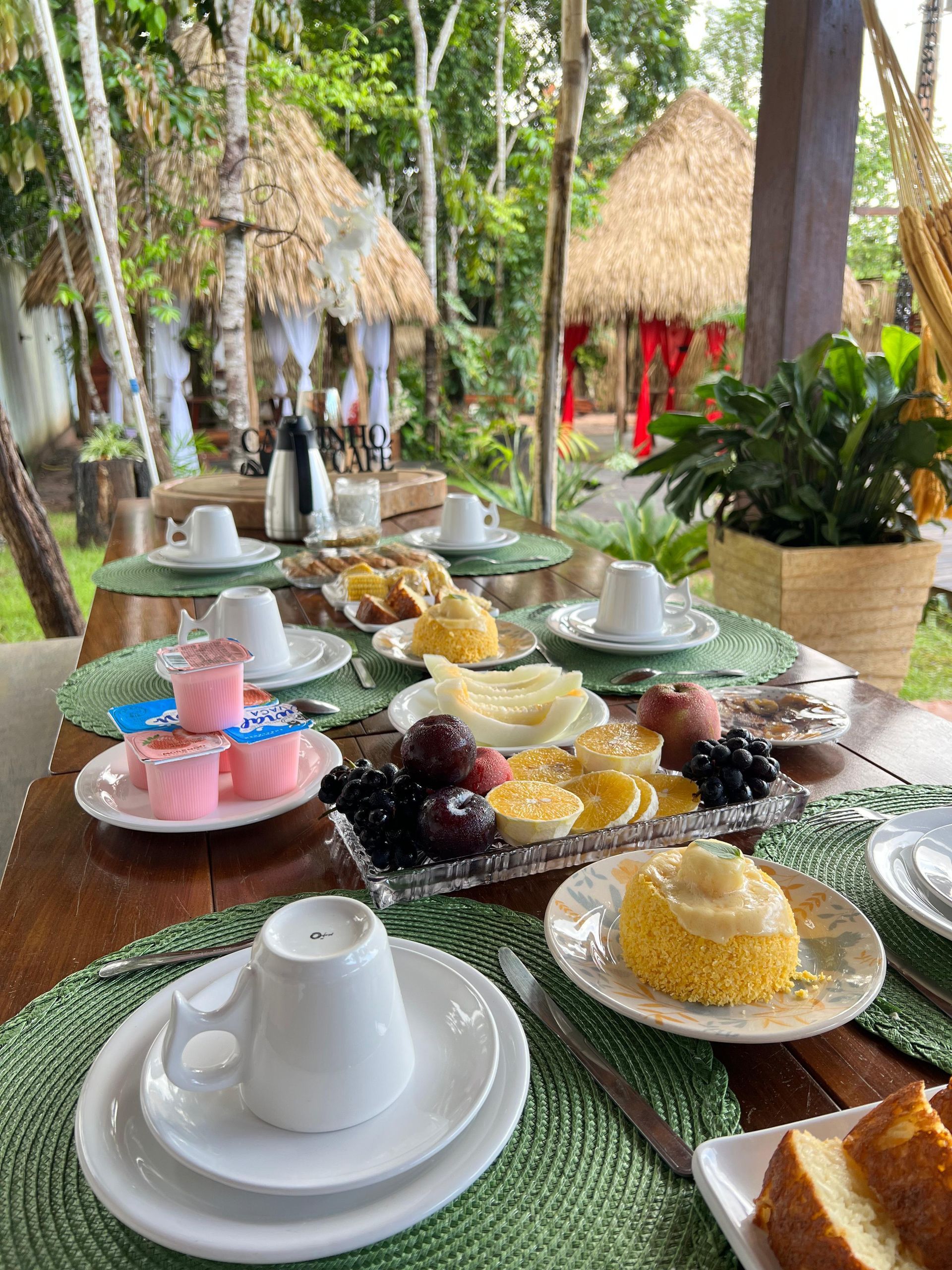 Café da manhã servido ao ar livre, com chalés de palha e decoração romântica ao fundo