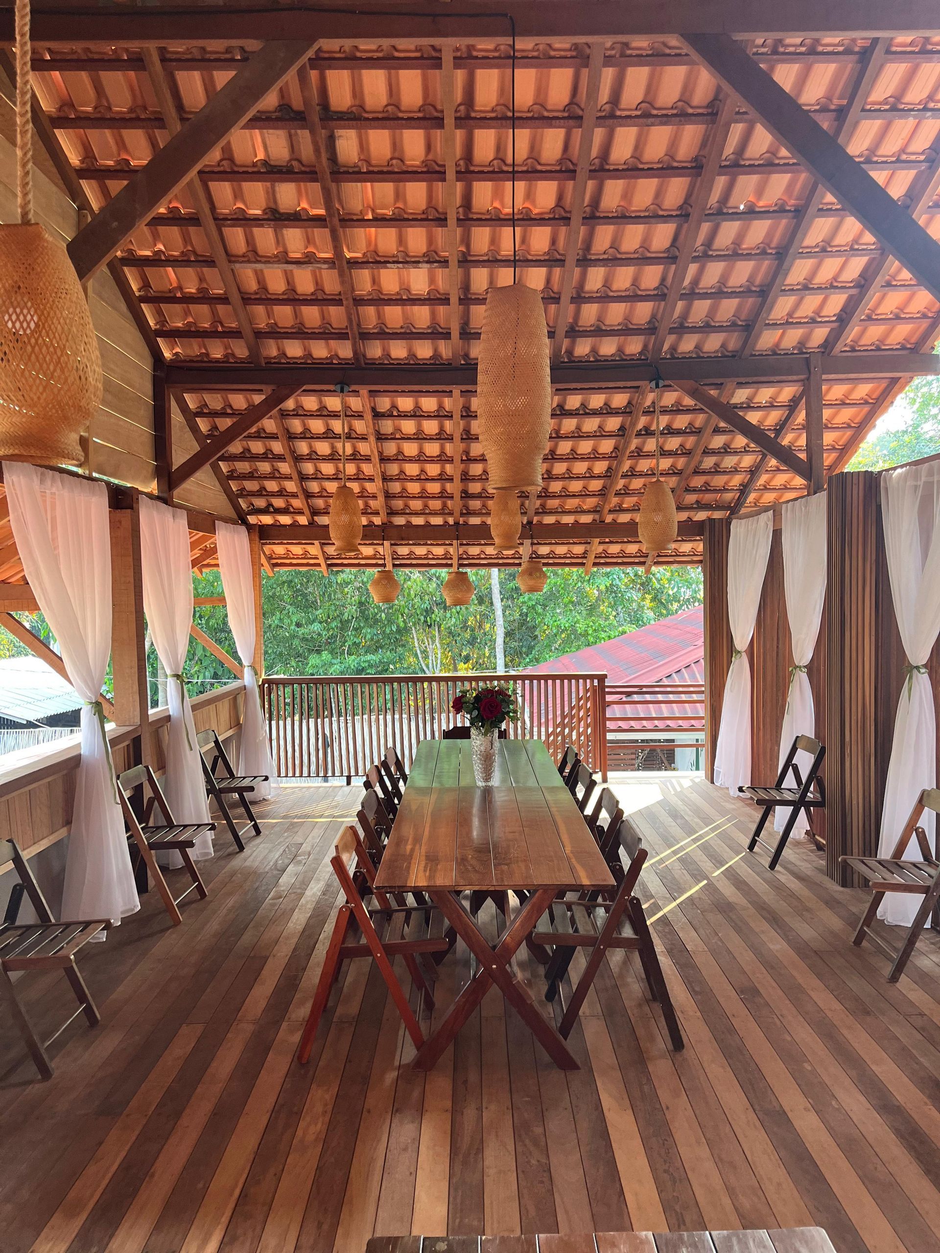 Vista frontal do deck de eventos com mesa longa, cadeiras dobráveis de madeira e luminárias de fibra