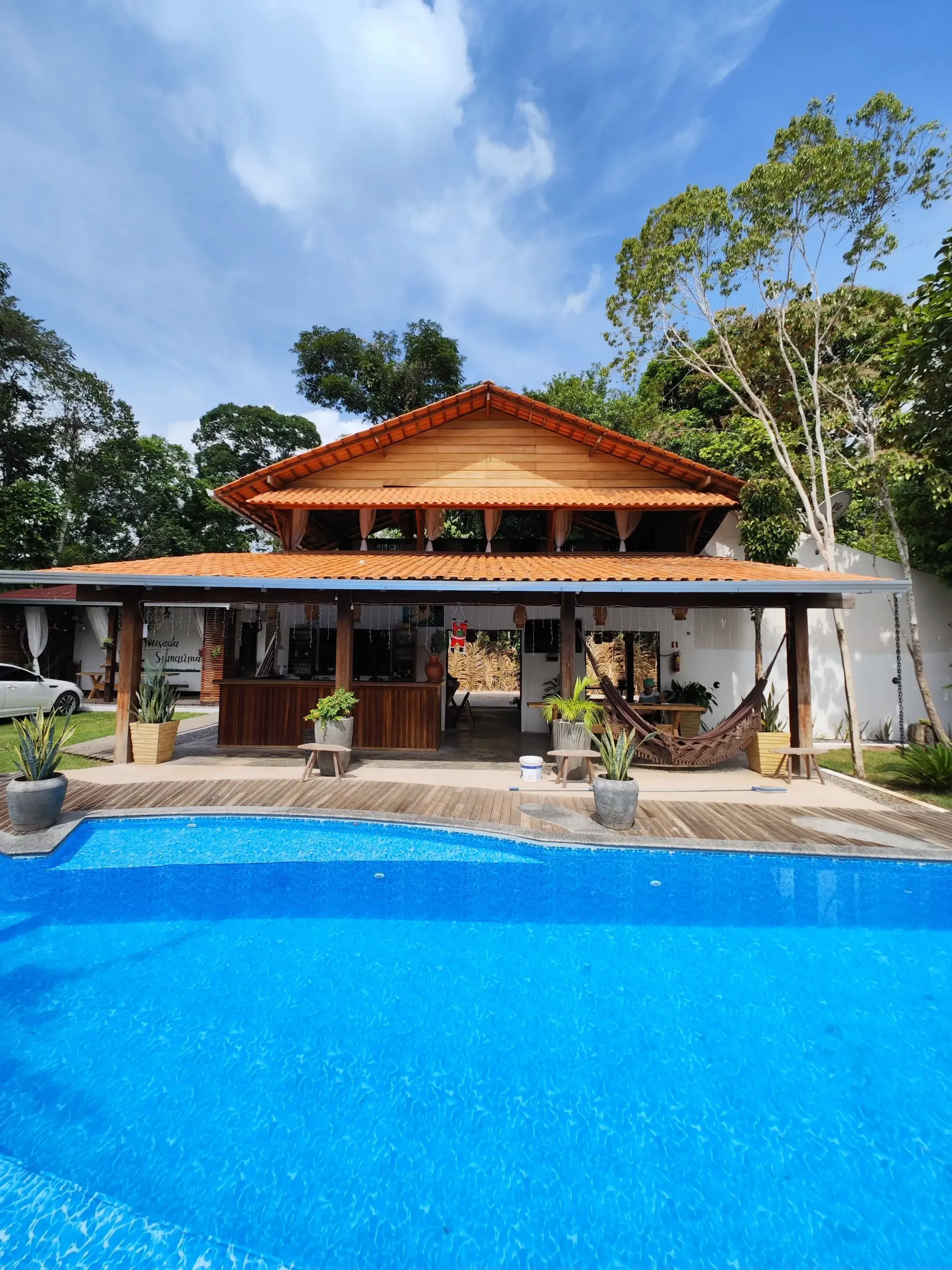 Piscina da Pousada Sumaúma diante do restaurante com telhado de madeira, cercada por árvores altas